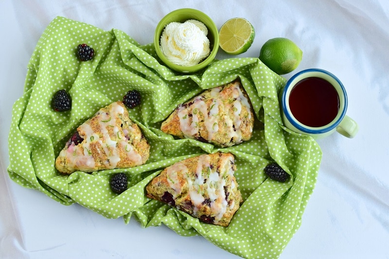 Three Blackberry Scones with Glaze on Green Cloth Surrounded by Blackberries, Cut Lime, Tea and Cream to Represent Blackberry Scone Recipe | Vitacost.com/Blog