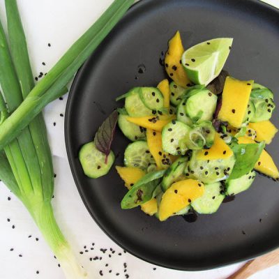 Thai Salad with Mango Chunks, Cucumber Slices, Scallions, Basil Leaves and Black Sesame Seeds on a Black Plate with a Bunch of Scallions Beside the Plate | Vitacost.com/blog
