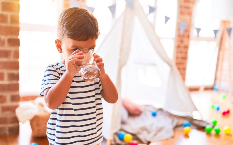 Concept of How to Get Kids to Drink Water Represented by Small Boy Sipping From Water Glass in Playroom | Vitacost.com/blog