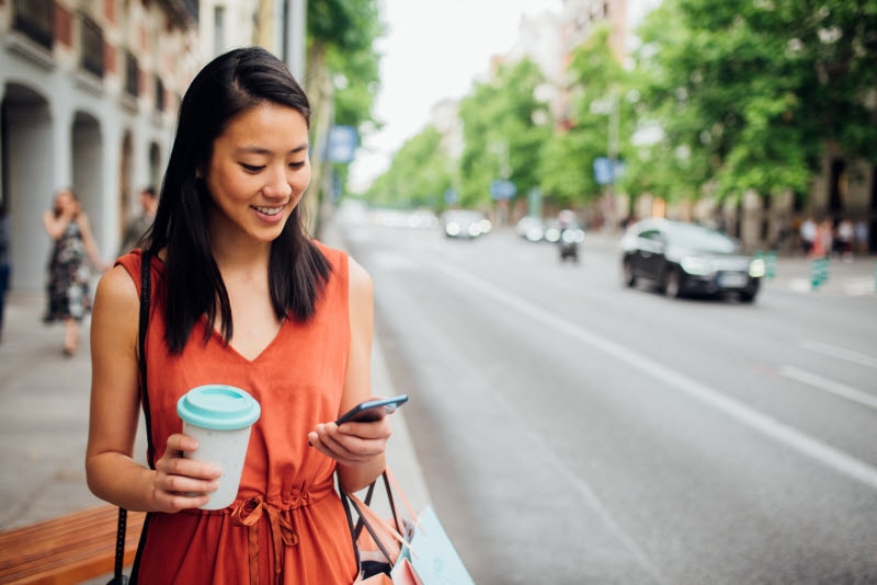 Woman Learning How to Improve Mental Health Walking Down Street Smiling and Looking at Phone | Vitacost.com/blog