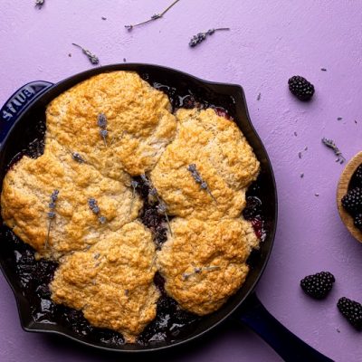 Blackberry Cobbler in Cast Iron Skillet Surrounded by Blackberries and Lavender Flowers on Light Purple Table | Vitacost.com/Blog