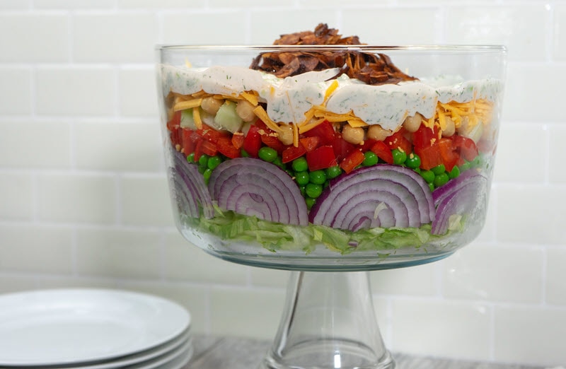 Salad in a Trifle Dish with Visible Layers of Lettuce, Red Onion, Peas, Red Bell Pepper, Vegan Shredded Cheese, Creamy Dressing Topped with Smoky Coconut Chips to Represent Layered Salad Recipe | Vitacost.com/blog