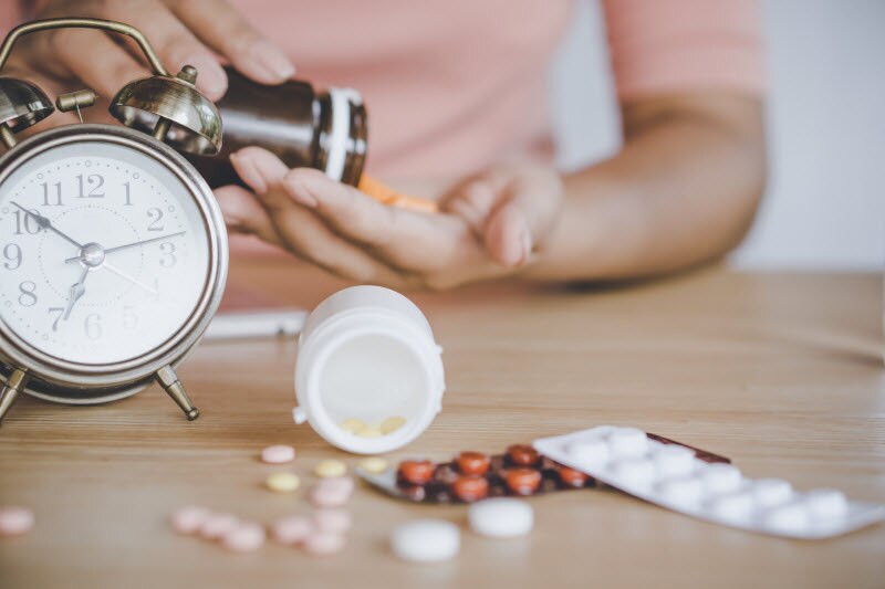 When is the Best Time to Take Probiotics Person Spilling Capsules from Bottle into Hand Beside Clock and Scattered Pills to Represent Concept of When it the Best Time to Take Probiotics | Vitacost.com/blog