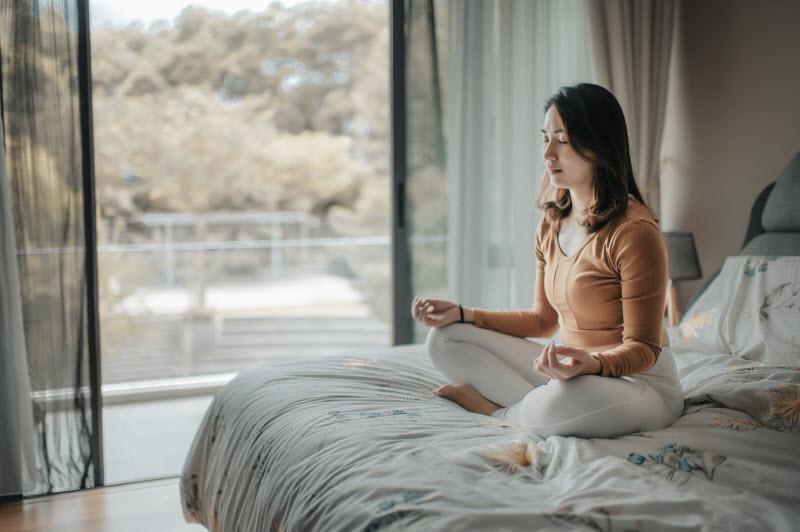 Woman Sitting in Bed at Dawn Trying Ayurvedic Practice of Meditation to Start Day Right | Vitacost.com/blog