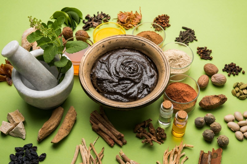 Herbs, Powders, Bowl with Mortar and Pestle on Green Surface to Represent Ayurvedic Medicine Essentials | Vitacost.com/blog