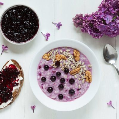 A Bowl of Purple Oatmeal With Berries, Nuts and Seeds Surrounded by a Bowl of Currant Jelly and Purple Flowers to Represent Black Currant Recipe | Vitacost.com/Blog
