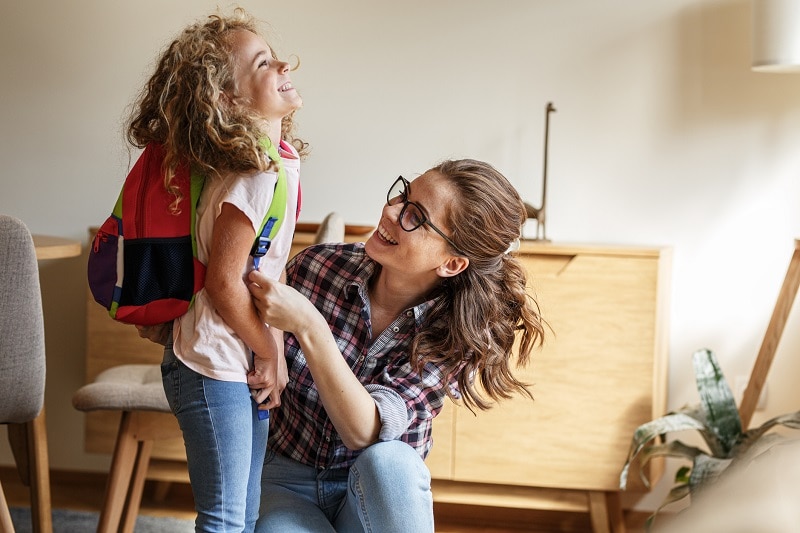 Smiling Mother Helps Young Smiling Daughter Put On Backpack to Represent Positive Attitude and School Anxiety | Vitacost.com/Blog