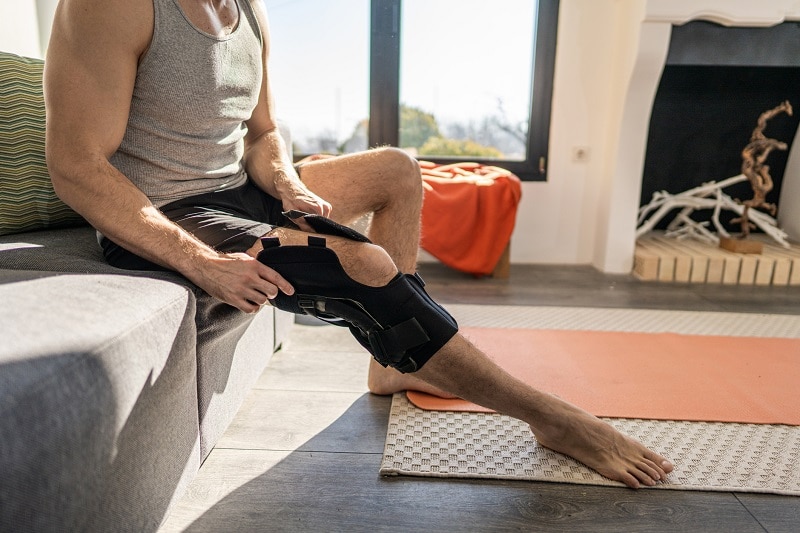 Young, Fit Man Sits on Sofa and Straps on a Knee Brace to Represent Supports and Braces | Vitacost.com/Blog