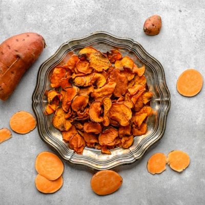 A Silver Plate Filled With Sweet Potato Chips is Surrounded by a Sweet Potato and Raw Sweet Potato Slices to Represent Air Fryer Sweet Potato Chips | Vitacost.com/Blog