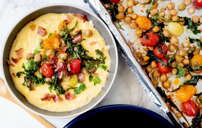 A Small Bowl of Grits is Topped With Roasted Arugula, Tomatoes, Chickpeas and Bacon Pieces Next to a Sheet Pan of Roasted Toppings to Represent a Cheese Grits Recipe | Vitacost.com/Blog