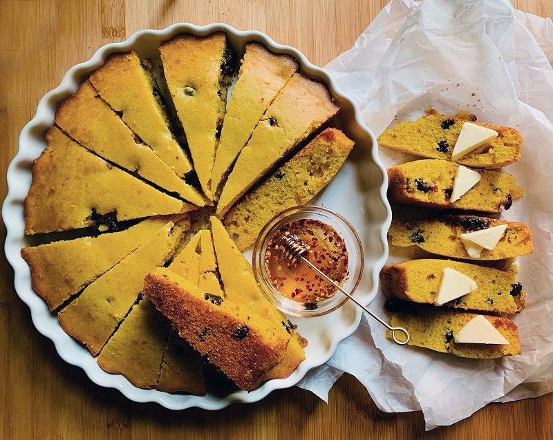 A Round Cornbread With Cranberries is Sliced into Wedges With a Small Bowl of Spicy Honey to Represent a Cornbread Recipe | Vitacost.com/Blog