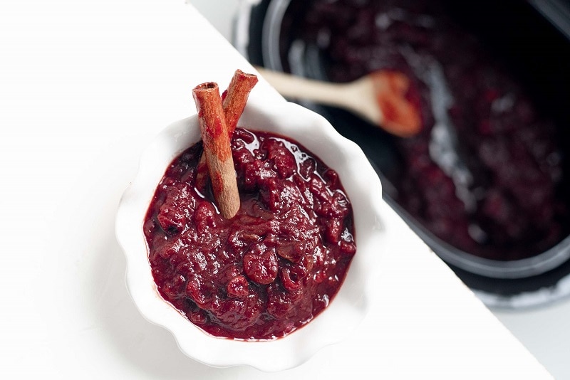 A Small White Bowl Filled With Slow Cooker Cranberry Sauce With Two Cinnamon Sticks and the Slow Cooker in the Background | Vitacost.com/Blog