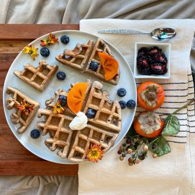 A Plate With Belgian-Style Waffles, Scattered Blueberries and Persimmon Slices Sits on a Wooden Tray to Represent a Gingerbread Waffle Recipe | Vitacost.com/Blog