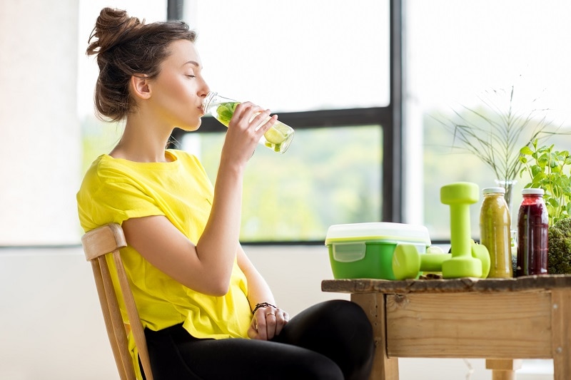 Young Woman Wearing Yellow Drinks Infused Water at a Table With Wellness Items to Represent a Detox Diet | Vitacost.com/Blog