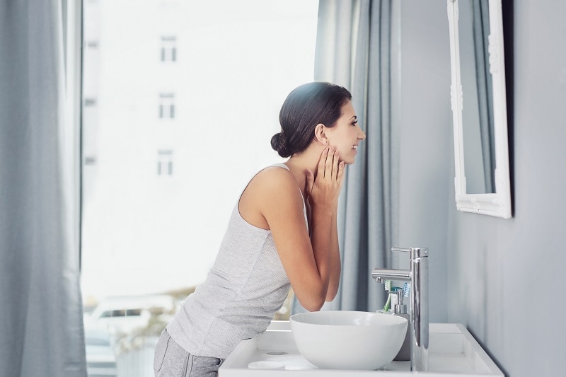 Beauty Goals 2022: Health & Happiness from the Inside Out A Woman Happily Looks in the Bathroom Mirror to Represent Beauty Goals