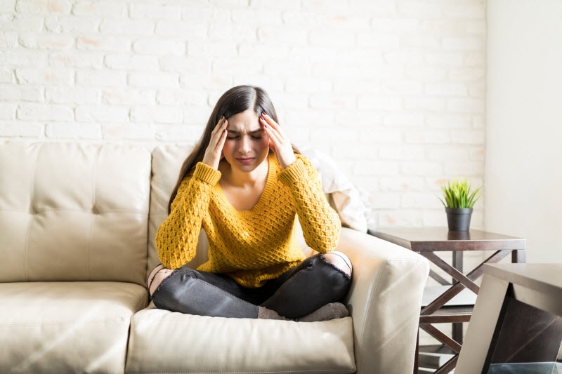 Woman Holding Head in Pain on Couch With Symptoms Magnesium Deficiency | Vitacost.com/blog