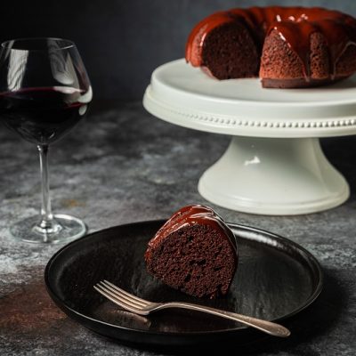 A Red Wine Chocolate Cake, Bundt Style, Sits Atop a White Cake Stand With One Slice on a Black Plate and a Glass of Red Wine Beside It | Vitacost.com/Blog
