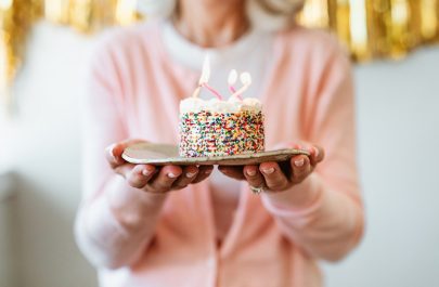 An Older Woman in Pink Shirt Holds a Plate With a Small Birthday Cake to Represent Healthy Aging | Vitacost.com/Blog