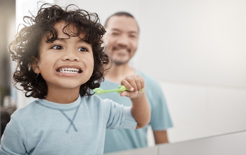 A Young Boy With Brown, Curly Hair Brushes His Teeth While His Father Looks On Smiling to Represent Children's Dental Health | Vitacost.com/Blog