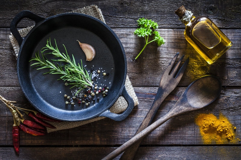How to Season a Cast Iron Pan Concept Represented by Overhead View of Pan with Herbs, Garlic and Oil on Wood Surface | Vitacost.com/blog