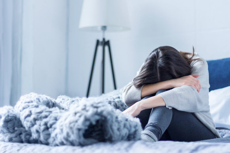 Woman With Anxiety About Health Sitting in Bed With Head Resting on Knees | Vitacost.com/blog