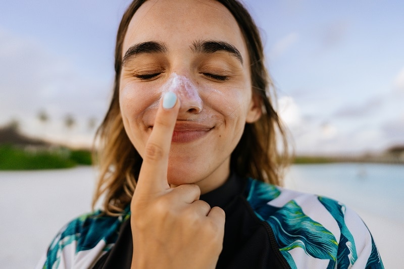 Chemical vs. Mineral Sunscreen – What’s Better? Woman Applying Mineral Sunscreen to Nose on the Beach | Vitacost.com/Blog