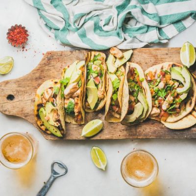 Chicken and avocado tacos in gluten-free shells on wooden serving board surrounded by limes and bowls of spices | Vitacost.com/blog