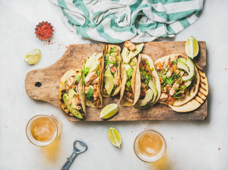 Chicken and avocado tacos in gluten-free shells on wooden serving board surrounded by limes and bowls of spices | Vitacost.com/blog