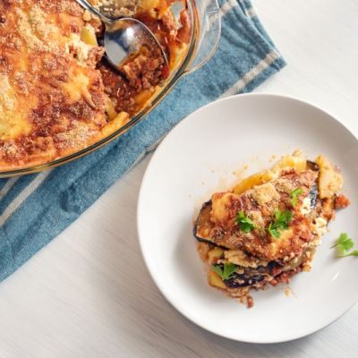 Greek moussaka casserole on white plate next to clear glass dish on blue napkin | Vitacost.com/blog