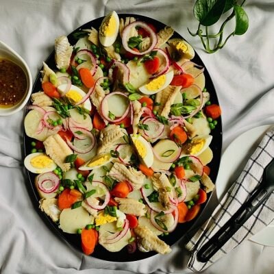 A Large Black Platter is Filled With a Bacalao Recipe of Root Vegetables Such as Carrots and Potatoes With Peas, Hard Boiled Eggs, Onions and More | Vitacost.com/Blog