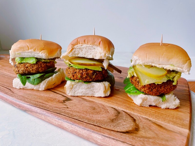 Three Homemade Black Bean Sliders Topped with Pickles, Onions, Lettuce and Avocado on Wooden Cutting Board | Vitacost.com/Blog