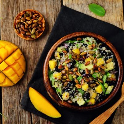 Quinoa, Mango & Black Bean Salad with Avocado, Arugula & Toasted Sunflower Seeds in Wooden Serving Bowl on Picnic Table Type Surface Next to Sliced Mango | Vitacost.com/Blog
