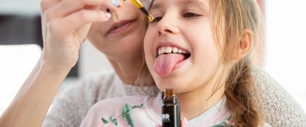 A Young Girl Sits on a Mother's Lap and Sticks Out her Tongue to Have a Dose of CBD for Kids | Vitacost.com/Blog