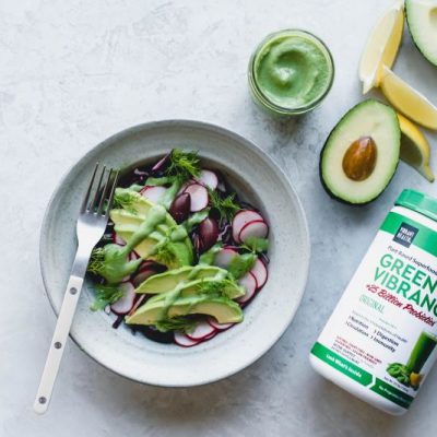 Vibrant Health Green Goddess Dressing on Salad in Bowl & in Glass Jar Next to Package of Greens Powder, Lemons & Avocados