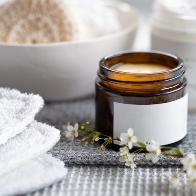 A Small Brown Glass Jar With Creamy Moisturizer is Surrounded With Bathroom Essentials, Such as Towels and Sponges to Represent a DIY Moisturizer.