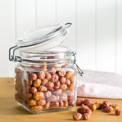 A Clear Jar Filled With Honey Roasted Nuts Sitting Open On A Counter With Additional Nuts Sitting On The Table