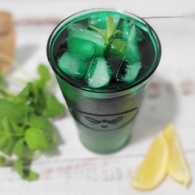 A Green Mint Chlorophyll Tea in Tall Glass With Ice. Lemon Slices and Mint Leaves Surround it on a Wooden Table