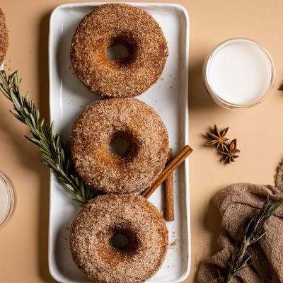 Three Baked Apple Cider Donuts Sit On a Long White Plate Surrounded By Star Anise, Small Bowls of Cream and Cinnamon Sticks.