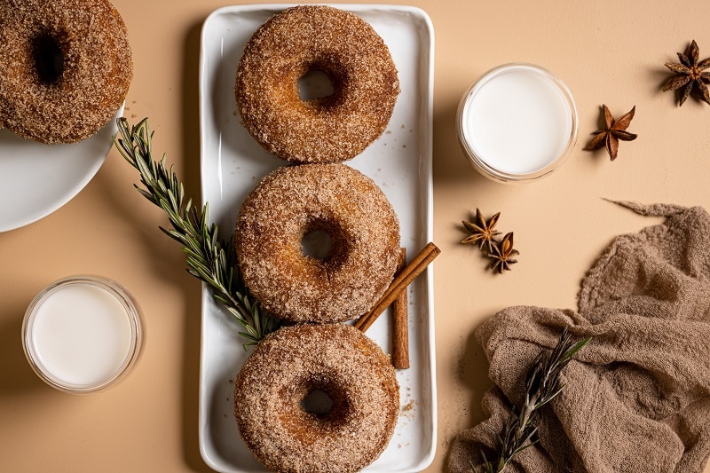 Three Baked Apple Cider Donuts Sit On a Long White Plate Surrounded By Star Anise, Small Bowls of Cream and Cinnamon Sticks.