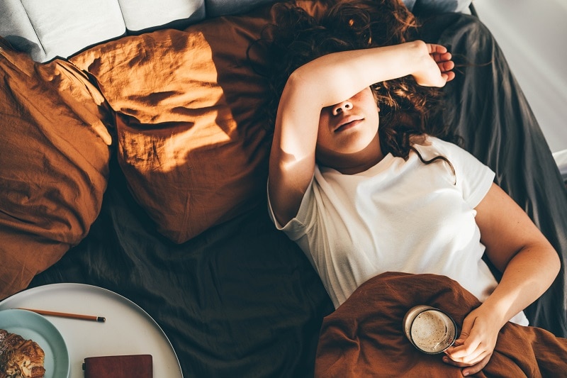 A Woman in Bed Covers Her Eyes With Her Arm While Holding a Cup of Coffee, Looking Sleep Deprived.