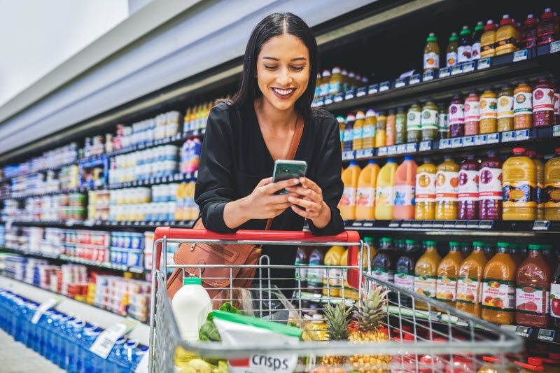 Woman Shopping in Grocery Store with Cart Checking Definition of Healthy on Her Phone