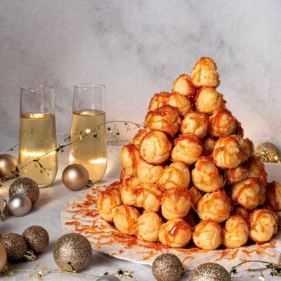French Croquembouche Puff Pastry Tower Drizzled in Caramel on Round Platter on Tablescape with Party Decorations and Glasses of Champagne