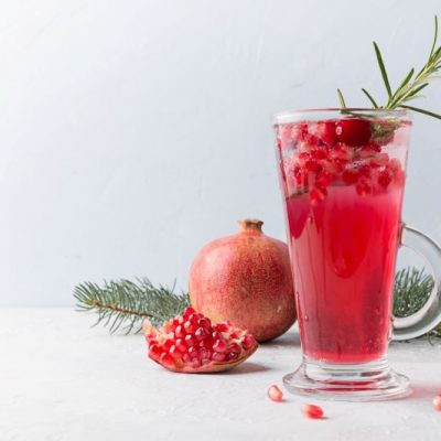 Pomegranate-Ginger Mocktail with Schisandra on Tall Glass Surrounded by Pomegranate & Greenery