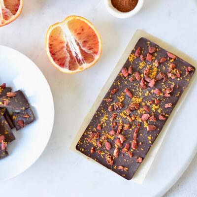 A White Plate With Pieces of Chili Chocolate Bark With Orange and Goji Berries Sits Beside Blood Orange Halves and a Whole Rectangle of the Bark.