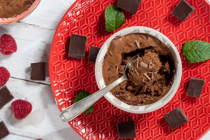 A Small Bowl is Filled With Chocolate Vegan Mousse, Sitting on a Bright Red Platter and Surrounded by Squares of Chocolate, Raspberries and Mint Sprigs.