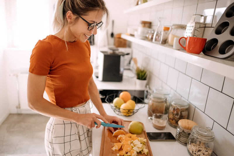 Smiling Woman Cutting Fruit in Kitchen to Represent Question is Organic Food Worth the Cost