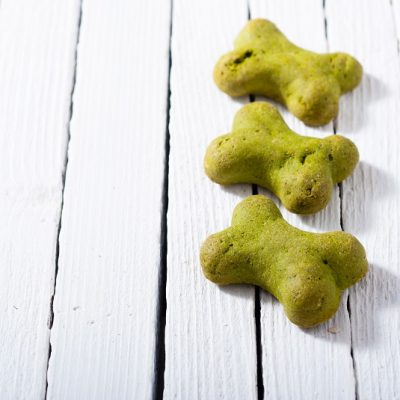 Three Homemade Green Dog Treats in the Shape of Bones are Placed Horizontally on a White Wooden Table.