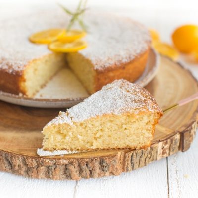 A Dusted Lemon Olive Oil Cake Has a Few Slices of Lemon and Sprig of Rosemary for Decoration With One Slice Cut and On Display.