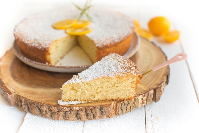 A Dusted Lemon Olive Oil Cake Has a Few Slices of Lemon and Sprig of Rosemary for Decoration With One Slice Cut and On Display.