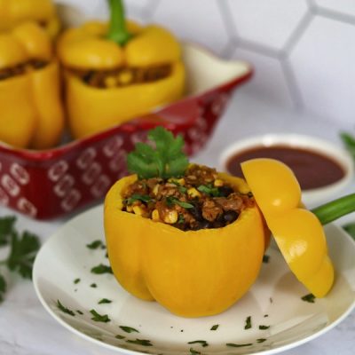 A Chicken Stuffed Pepper Sits With a Garnish of Cilantro in Front of a Larger Serving Platter of Chicken Stuffed Peppers.